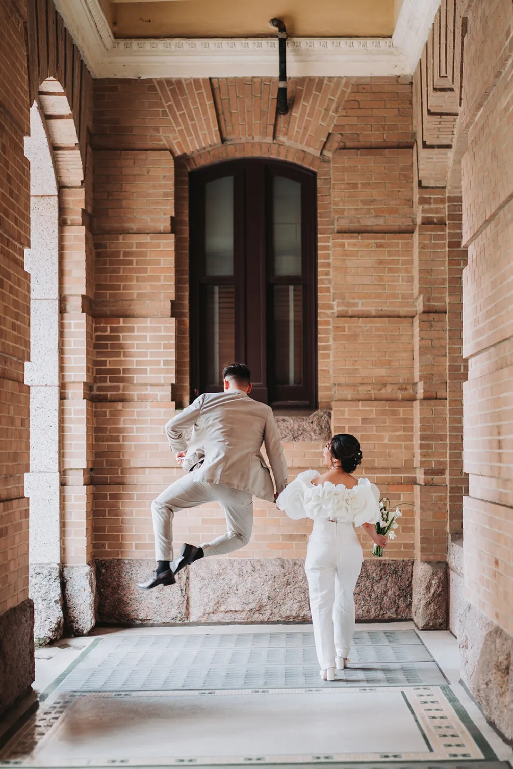 13 engagement photos in dallas houston texas A joyful bride and groom in elegant attire are walking hand in hand along a brick corridor, capturing a lively moment as the groom leaps into the air in celebration.